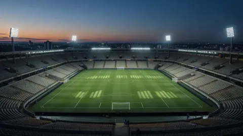 Vista aérea de um grande estádio de futebol ao entardecer, com arquibancadas vazias e refletores acesos.