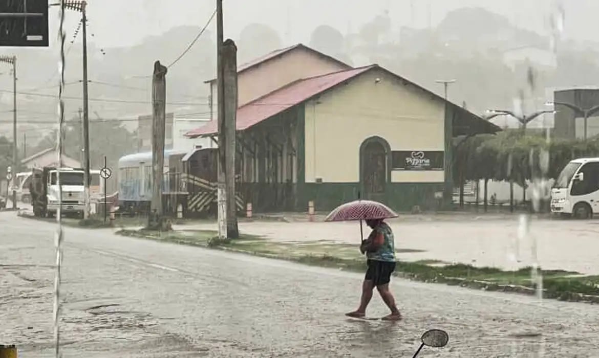 Pedestre com guarda-chuva atravessa rua alagada durante chuva forte em área urbana.