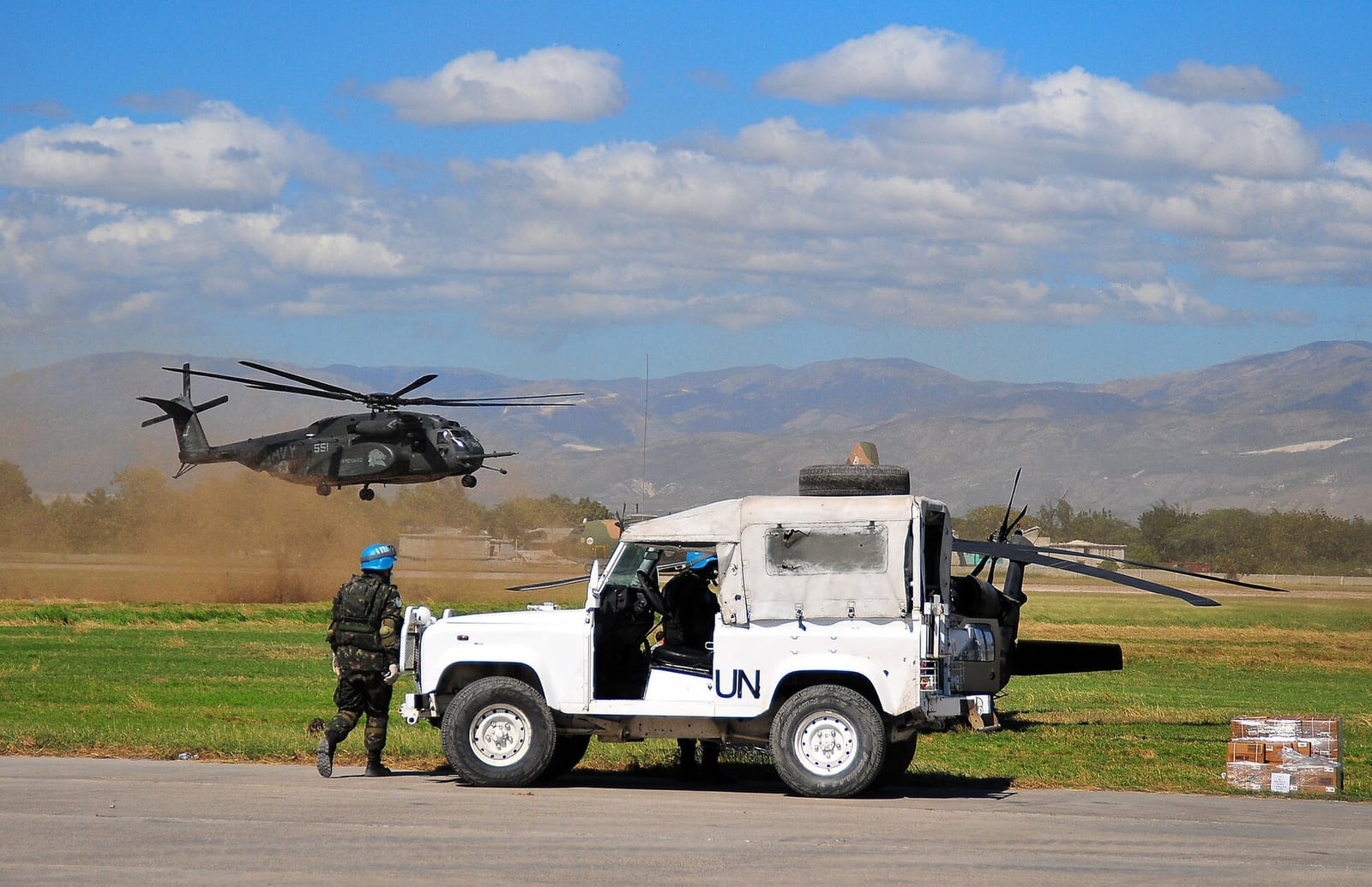 Helicóptero militar sobrevoa veículo da ONU enquanto um integrante de capacete azul se aproxima, em área aberta no Haiti.