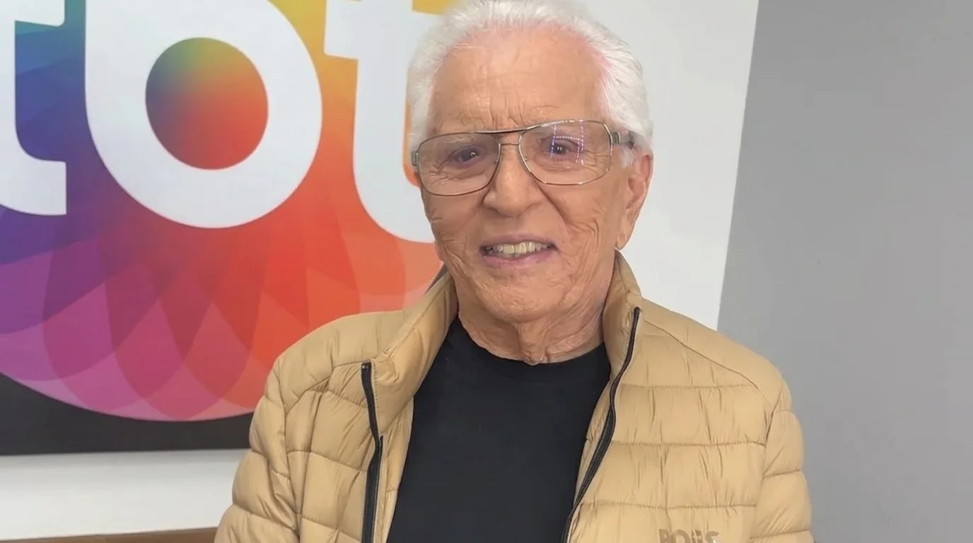 Carlos Alberto de Nóbrega, de jaqueta bege e óculos, posa sorrindo diante de um painel colorido.