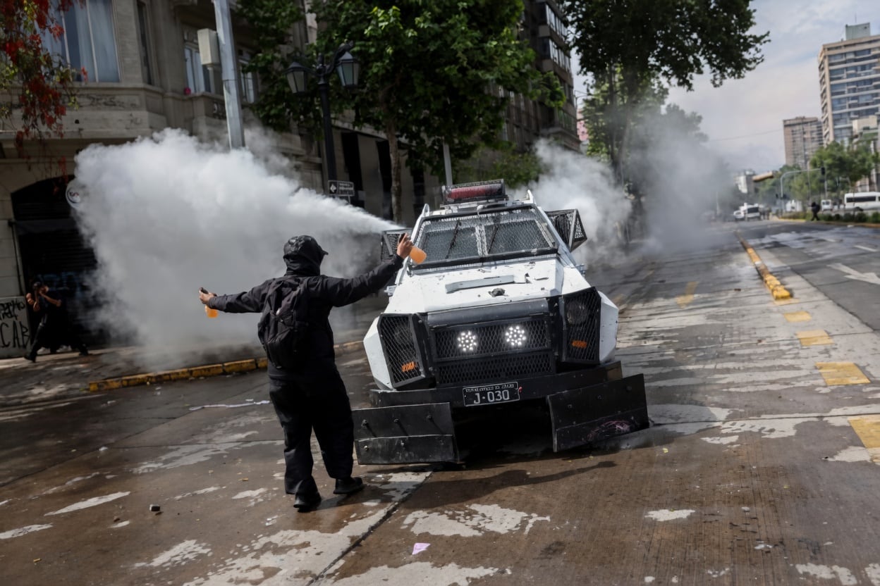 Manifestante mascarado ao lado de um veículo blindado da polícia, em meio a fumaça branca em uma via urbana.