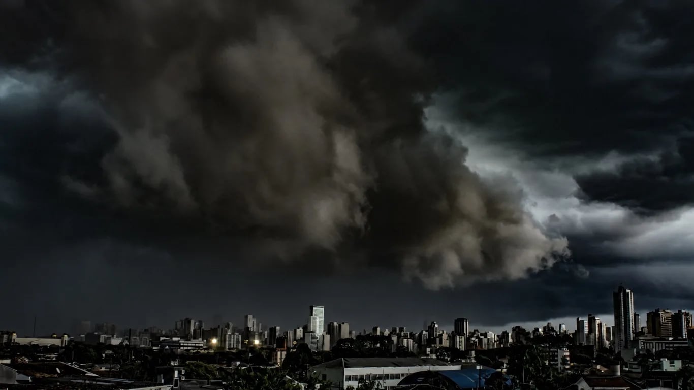 Nuvens escuras de chuva forte cobrem o céu sobre prédios e casas de uma área urbana.