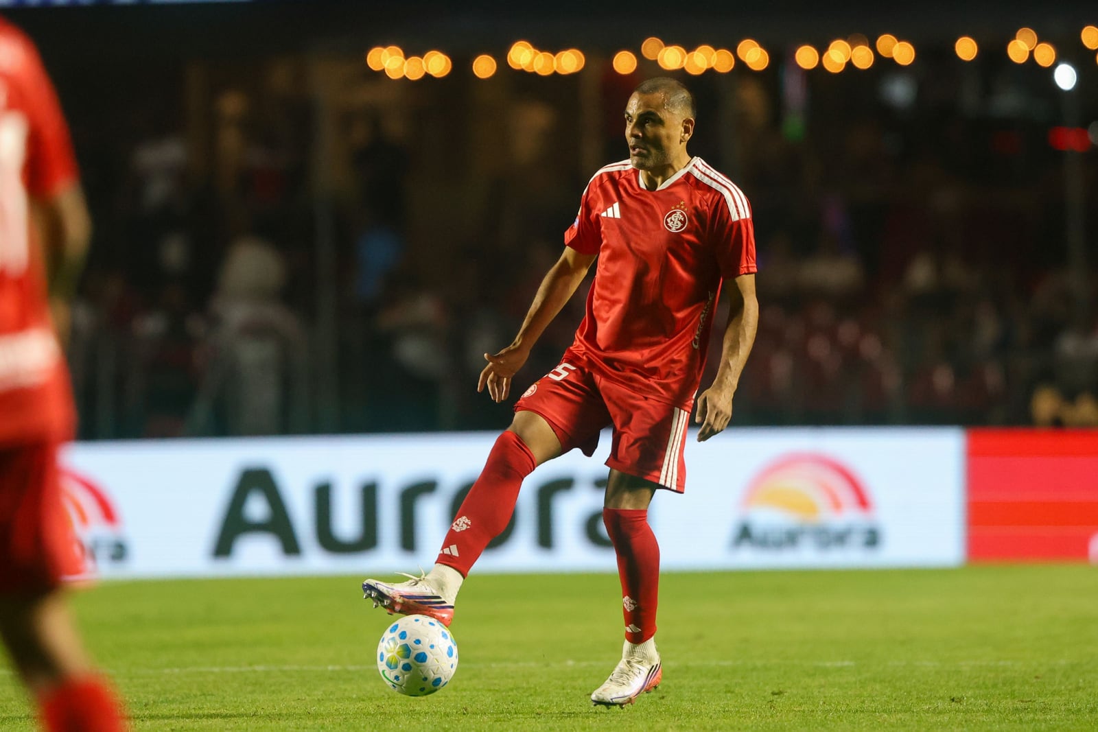 Jogador do Inter, de camisa vermelha, conduz a bola durante partida, com placa publicitária da Aurora ao fundo do gramado.