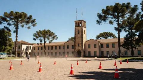 Prédio público de estilo quartel, de paredes bege e torre de pedra com mastros de bandeira, sob céu azul e