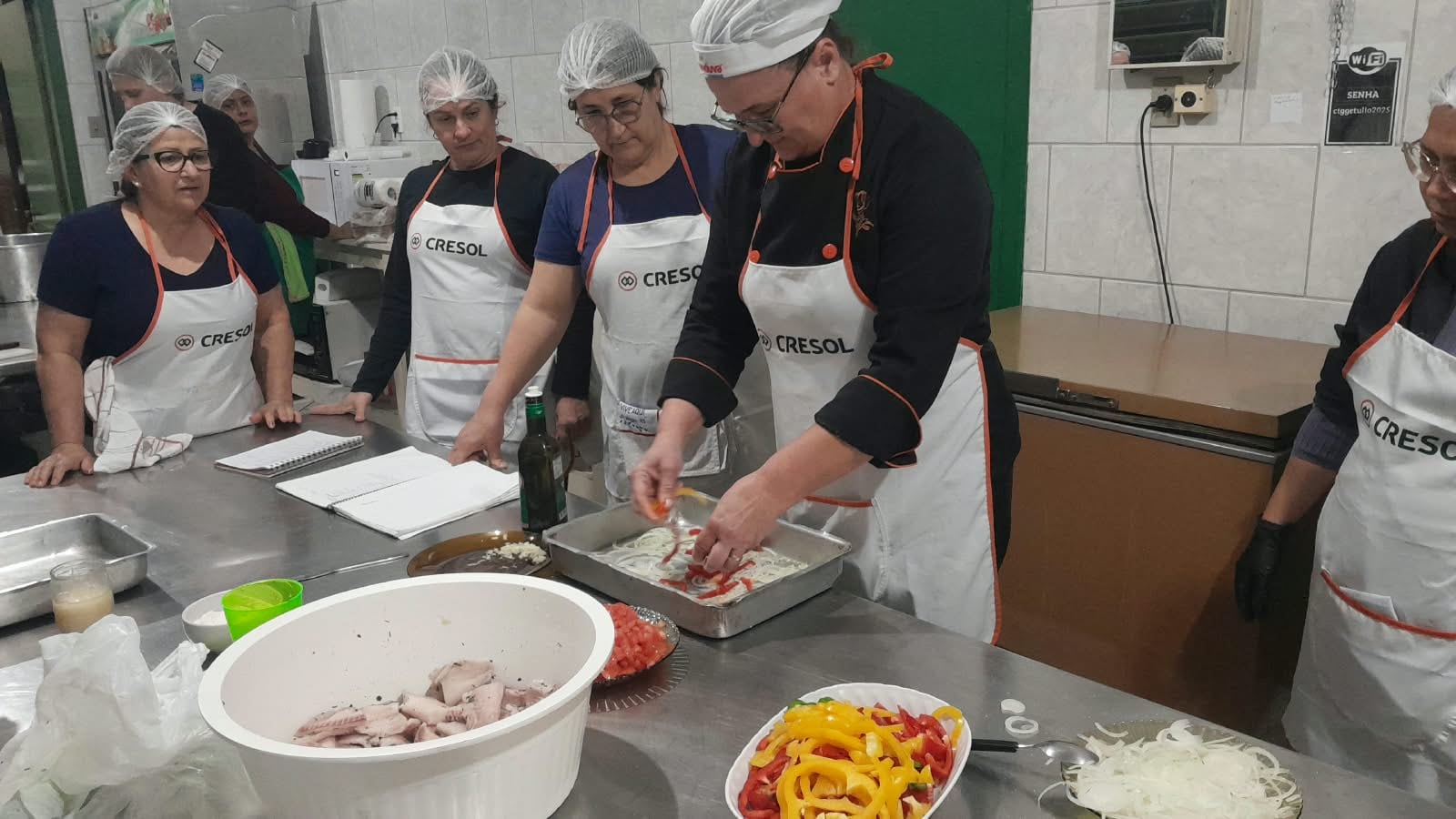 Aula prática do curso Arte de Cozinhar, com instrutor preparando alimentos em cozinha industrial e alunos de avental observando.