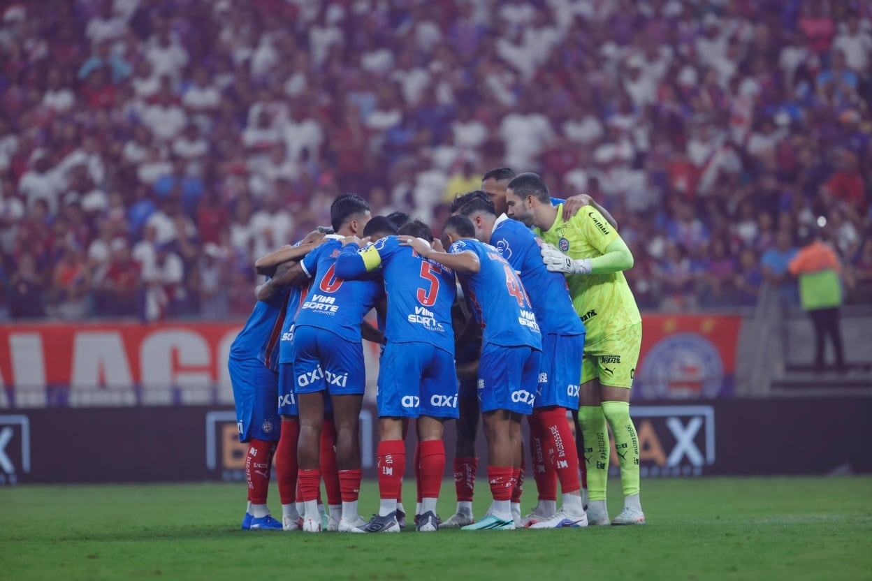 Jogadores do Esporte Clube Bahia reunidos em roda antes de uma partida, com torcida ao fundo.