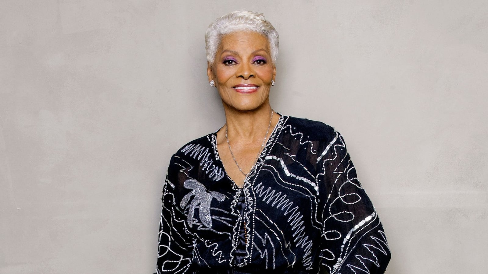 A cantora Dionne Warwick posa com blusa preta bordada em prata, diante de um fundo cinza.