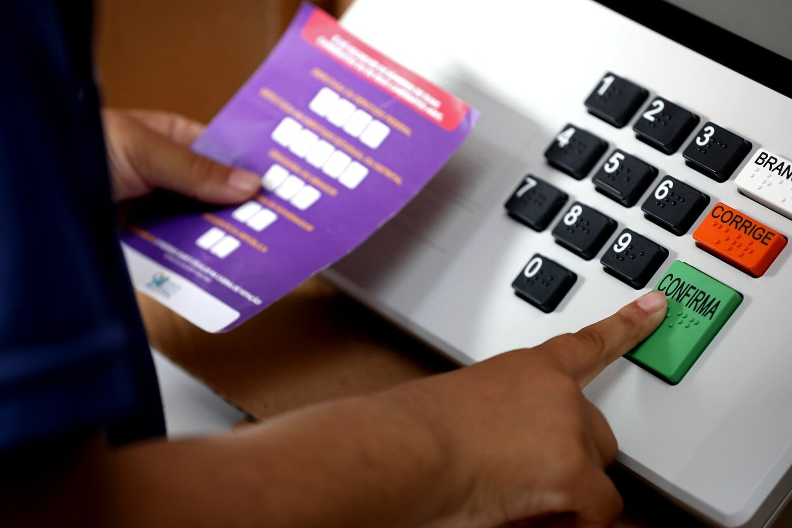Mão de uma pessoa pressiona a tecla CONFIRMA de uma urna eletrônica, ao lado de um papel com instruções de votação.