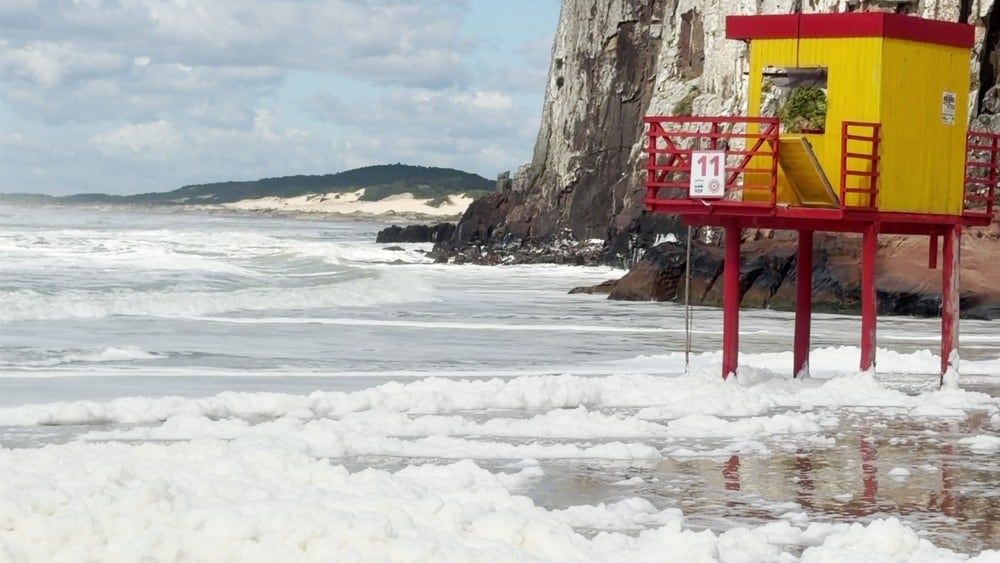 Espuma branca se espalha na faixa de areia da praia de Torres, ao lado de uma torre de salva-vidas.