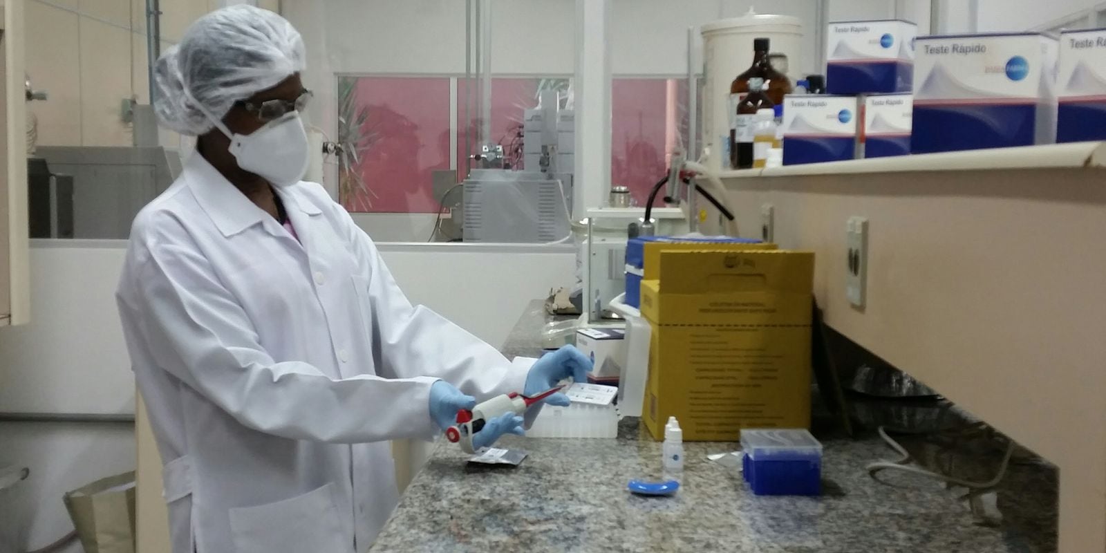 Profissional de jaleco, touca e máscara manuseia equipamento em bancada de laboratório.