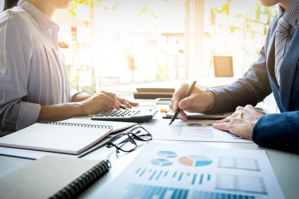 Duas pessoas analisam documentos com gráficos e uma calculadora sobre a mesa, em cena de trabalho financeiro.