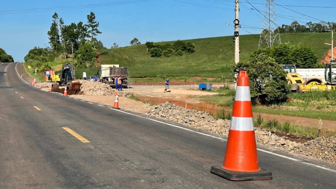 Trecho de rodovia com obras de terraplanagem, máquinas, caminhão e cones de sinalização ao lado da pista.