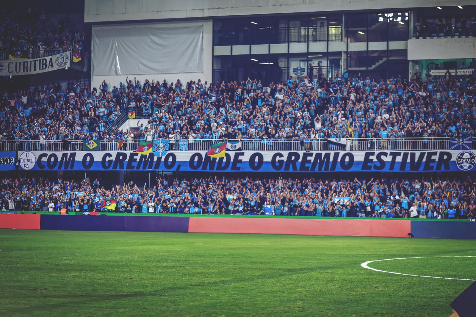 Torcedores do Grêmio na arquibancada, com faixa "Com o Grêmio, onde o Grêmio estiver".