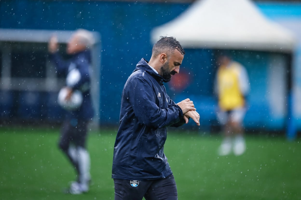 Homem com jaqueta azul do Grêmio consulta o relógio durante treino sob chuva, com o gramado molhado ao fundo.