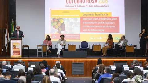 Mulher negra de blazer fala ao microfone em evento com painel e público sentado, durante lançamento de publicação sobre