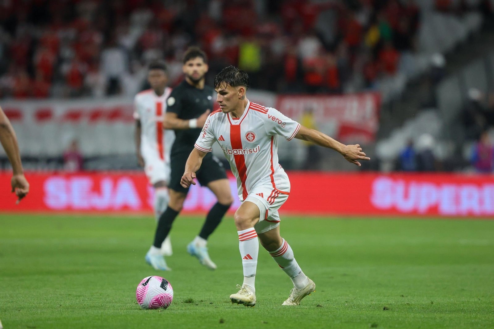 Jogador do Inter conduz a bola em partida com uniforme branco, sob o olhar de adversário ao fundo.