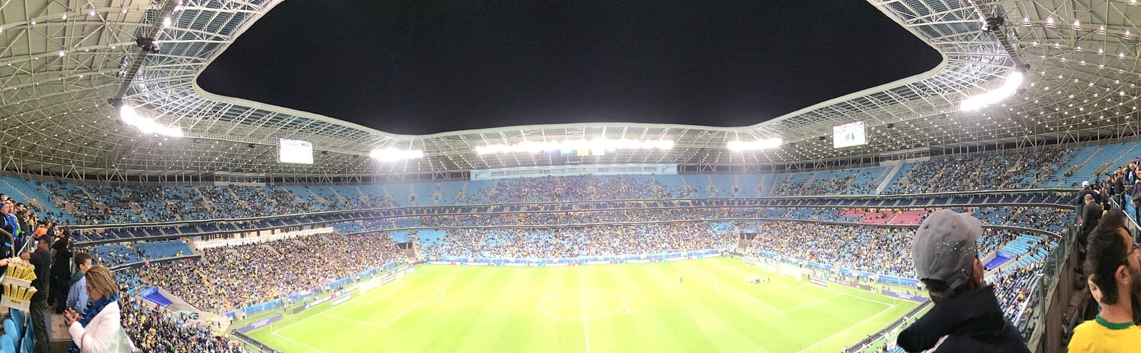 Vista panorâmica da Arena do Grêmio durante uma partida, com o gramado e as arquibancadas cheias.