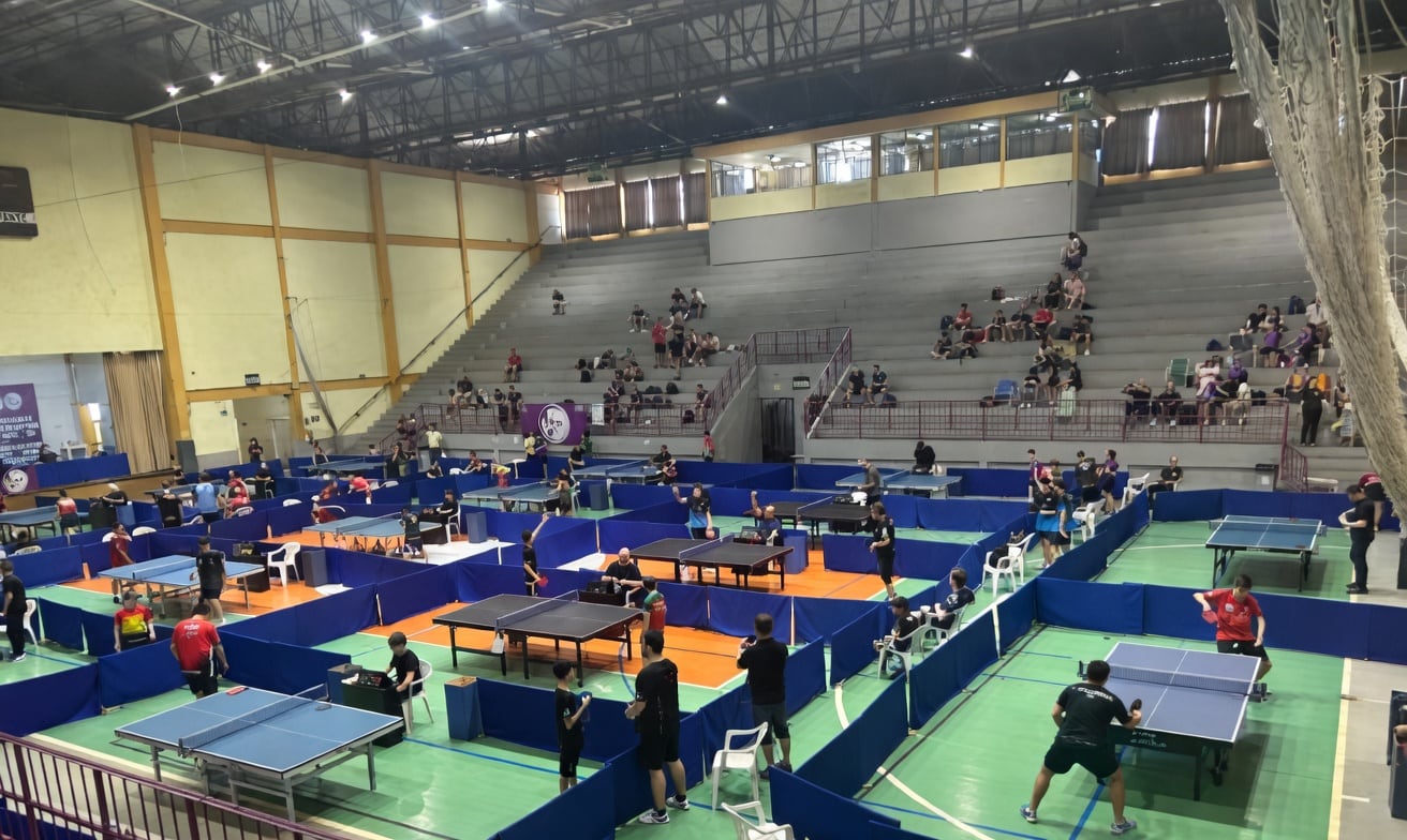Vista geral de um ginásio com mesas de tênis de mesa montadas, atletas em jogo e arquibancadas ao fundo.