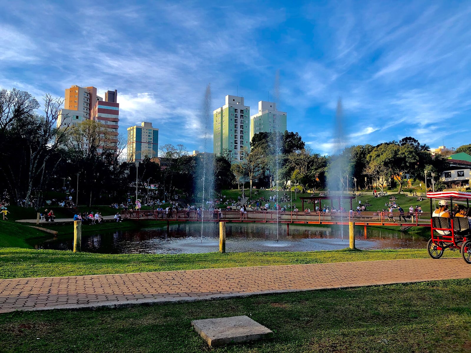 Vista do Parque da Gare, em Passo Fundo, com lago, chafarizes e pessoas passeando.
