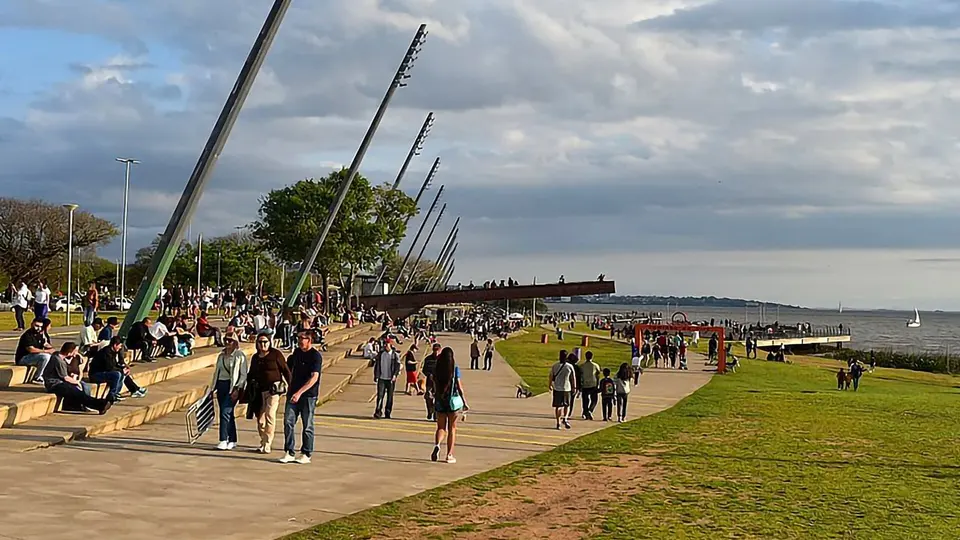 Dia ensolarado com céu azul sobre a orla de Porto Alegre, com pessoas caminhando e árvores ao redor