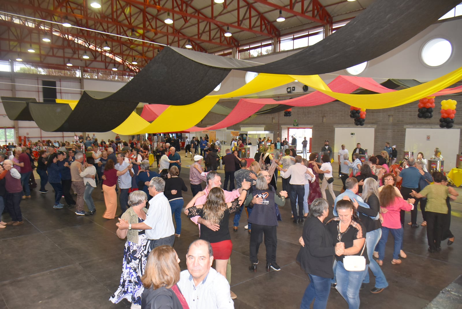 Público dança em salão decorado com faixas nas cores da bandeira alemã durante a Oktoberfest Erechim 2025.