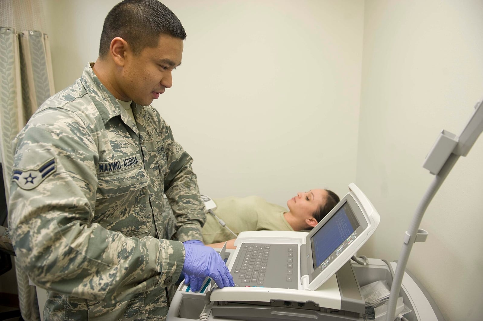Profissional de saúde da Força Aérea dos EUA opera equipamento de exame cardiológico em uma clínica, com uma paciente deitada ao fundo.
