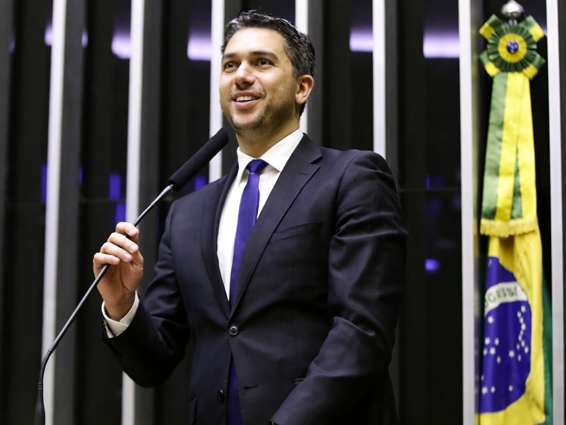 O deputado federal Daniel Trzeciak discursa ao microfone, com a bandeira do Brasil ao fundo.