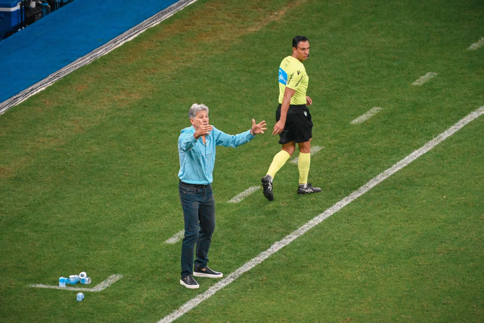 Renato Gaúcho gesticula à beira do campo durante uma partida, ao lado de um árbitro.