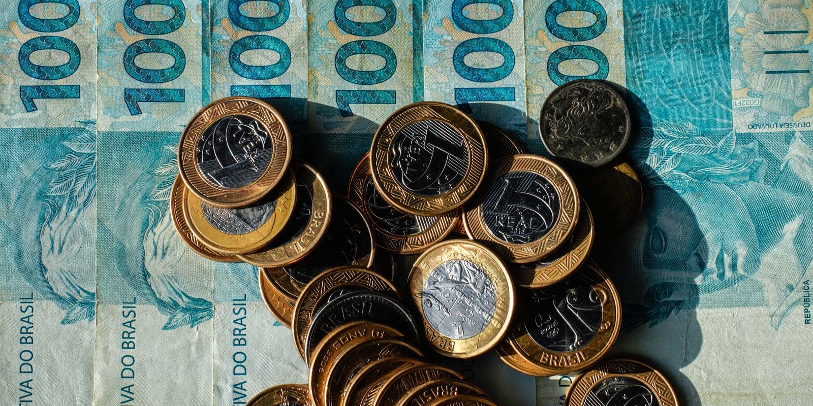 Moedas e cédulas de R$ 100 espalhadas sobre a mesa.