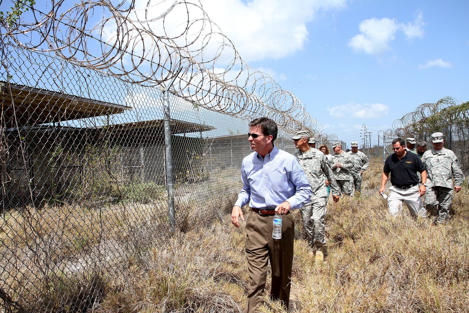 Ex-comandante da prisão de Guantánamo visita o complexo de detenção, cercado por cercas de arame farpado, acompanhado de militares.
