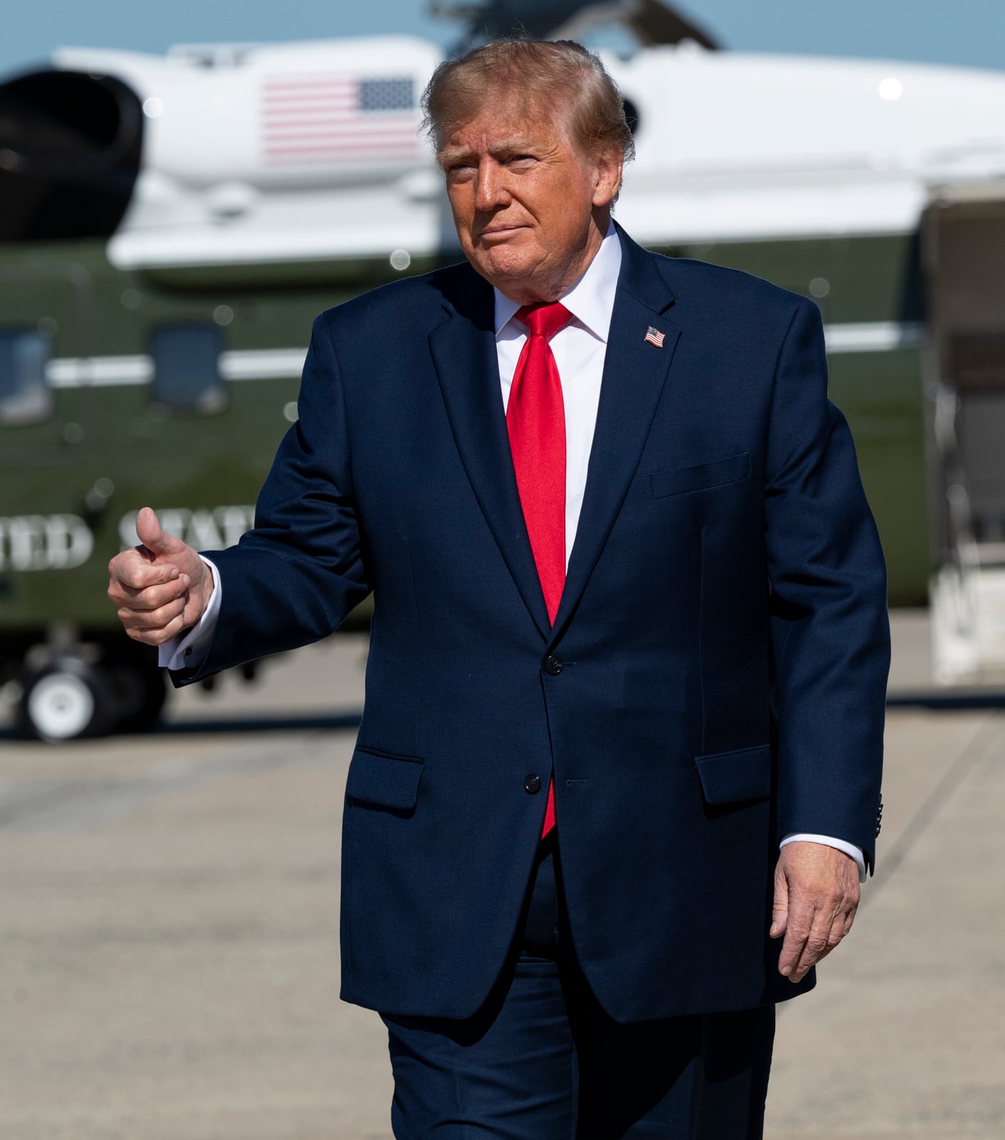 Donald Trump caminha em área de embarque, com helicóptero presidencial ao fundo.