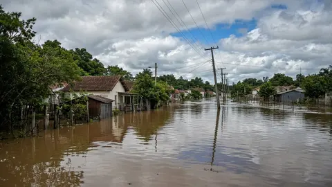 Ilustração: rua residencial alagada com água barrenta cobrindo o asfalto e os quintais, casas e galpões pela metade, céu